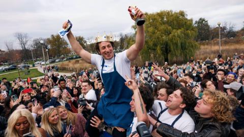 Ben Shabad, the winner of the Jeremy Allen White lookalike contest, wearing a chef's apron and white t-shirt, is hoisted on the shoulders of crowd members in a Chicago park