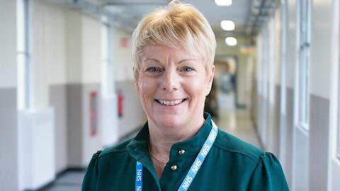 Michelle Arrowsmith, a woman with blonde short hair standing in a hospital corridor. She is looking directly at the camera and smiling and is wearing a dark green blouse and blue NHS lanyard round her neck.
