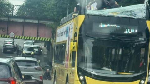 People are standing on the top deck of a yellow bus which has had much of its roof ripped off. The front windscreen has been smashed. 