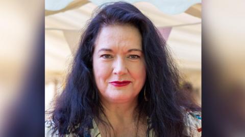 Image shows Ann Millington looking at the camera, she has long dark hair and is wearing a floral top