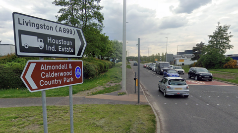 Houstoun Road in Livingston. Industrial units sit on the left with cars stopped at traffic lights on the road.