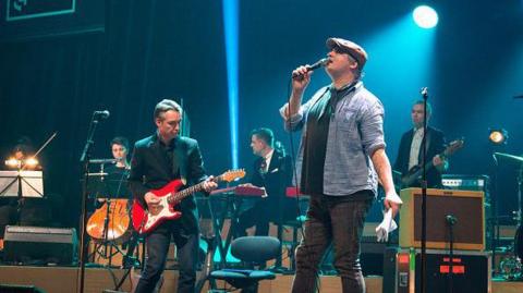 Members of a band perform on stage. The lead singrer wears a flat cap, denin shirt and black T-shirt and jeans and is holding a microphone. A man next to him is playing a red and white electric guitar, dressed in a black suit jacket, shirt and jeans. Behind them are three other musicians playing various instruments on the stage and dressed in suits. There are various speakers and microphones on the stage too.