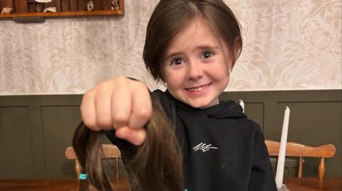 Reuben smiles at the camera while holding one arm out in front of him as he holds on to his hair that was cut. He wears a black hoodie and has brown hair.