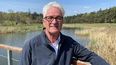 Melvin Benn has white hair and black glasses, and is wearing a dark grey jacket with a white shirt underneath. He's leaning against a wooden railing with a lake, reeds and trees behind him. It's a sunny day and the sky is blue.
