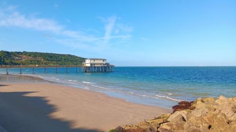 A short pier can be seen coming out over the water with a small white building on it. In the foreground, is a sandy beach with some rocks on the right. Both the sea and sky are bright blue. There is a hill covered in trees in the background.