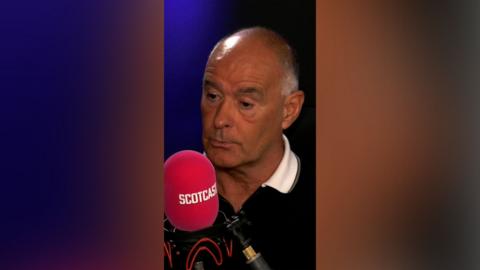 Tommy Sheridan on Scotcast wearing a dark polo shirt with white collar. He is speaking into a pink microphone with the word 'Scotcast' on it.