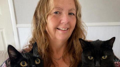 A middle-aged woman with blonde hair smiles at the camera. Two black cats are visible in front of her.