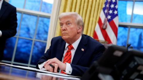 Donald Trump sits behind a desk at the White House holding a pen