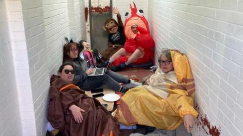 A group of five people wearing inflatable animal costumes - including a lobster and a monkey costume - sit on the floor of a brick kennel at animal shelter.
