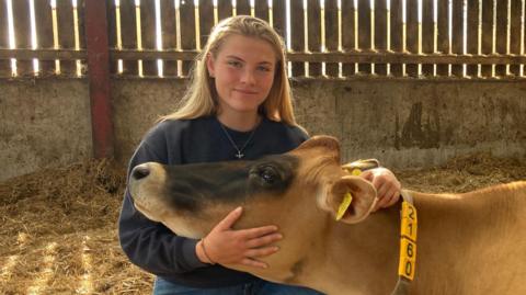 Erin Gailey in a barn holding a cow's head. She has long, light hair past her shoulders and wears a blue top with a gold necklace and blue jeans as she kneels on straw in an enclosed building. The cow has brown fur apart from darker fur on its nose and a plastic necklace with numbers on it around its neck and a yellow tag in its ear