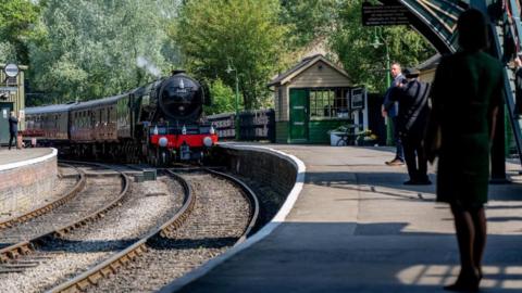A steam locomotive pulling into a historic-looking railway station platform. The station name of Pickering is just visible on a sign, and there are four people standing on the platform.
