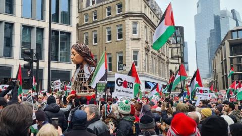 Little Amal, a 3.5 metre tall puppet, walking alongside protesters carrying Palestinian flags. Placards with "Gaza: stop the massacre" and "From the river to the sea Palestine will be Free" can also bee seen.