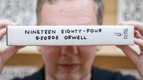 Artist David Shrigley holding a copy of 1984 by George Orwell over his eyes..