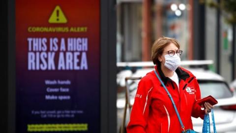 A woman wearing a protective mask walks past a warning sign in Greater Manchester