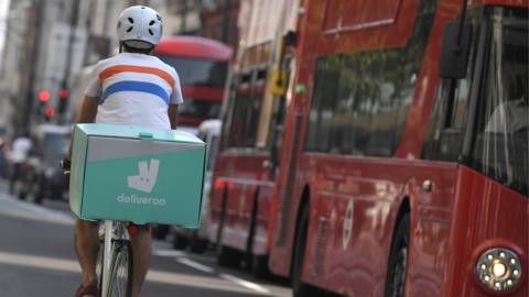 A bicycle courier with a box bearing the logo of Deliveroo rides through the streets of London with the city's iconic red buses visible nearby