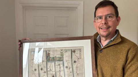 David Mitchell from Minety looks at the camera wearing glasses and a woolly jumper holding the framed map