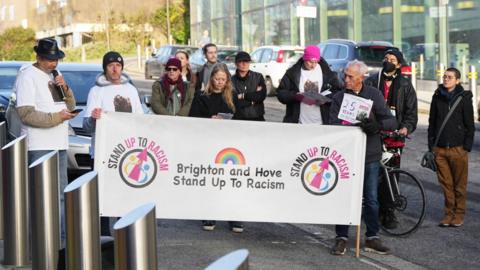 A group of people wearing t-shirts showing the face of Jay Abatan and carrying a banner which reads Brighton and Hove stand up to racism