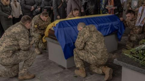 Ukrainian soldiers kneel before a coffin covered with a Ukrainian flag