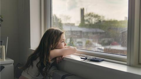 A young girl stares out of a window on a rainy day, looking unhappy