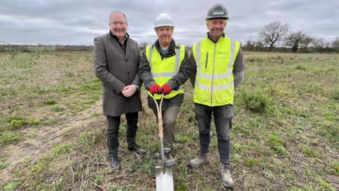 Three people stood in a field with a spade, marking the start of construction of the Maldon crematorium.