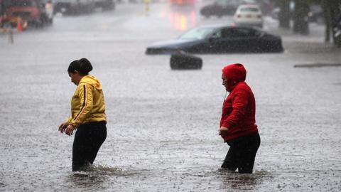 Residents walk through floodwaters in New York, with a submerged car in background