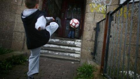 Two young boys play football in a run down street with boarded up houses