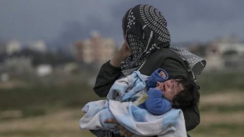 A Palestinian woman carries her baby as smoke rises below her in the Gaza Strip. Photo: 26 February 2024