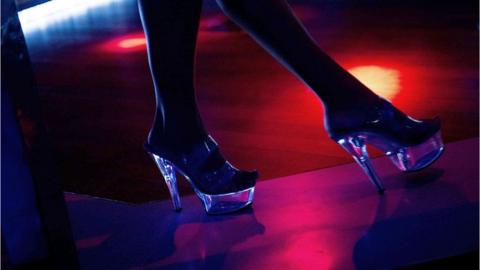 Image showing a silhouette of a woman's legs and high heels One foot is in front of the other. It is a dark image with red lighting. The heels are reflecting the light.