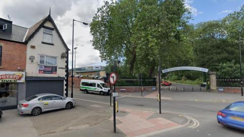 The junction of Barnsley Road and Herries Road in Sheffield. On the left of the junction there is a takeaway with a silver taxi parked outside. Across the road there is an arched entrance to Northern General Hospital.