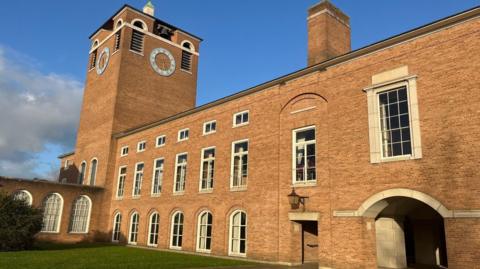 An exterior view of an L-shaped part County Hall in Exeter. It is a neoclassical-style brick building with a clock tower.