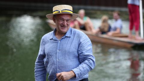 A man wearing a light blue shirt and a beige straw hat. He is standing on a boat on a river and is looking to his left and smiling. Behind him is a punt with people sitting on it.