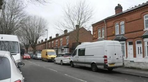 A residential street lined with terraced houses and trees. Cars and vans are parked both sides of the road.