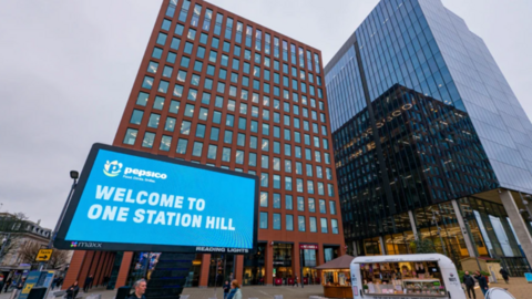 The view of two tall buildings from the ground behind a large sign saying 'Welcome to One Station Hill'