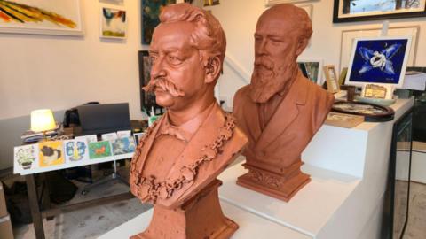 Two brown-coloured busts depicting two men. They have been placed on a white platform inside an art studio.