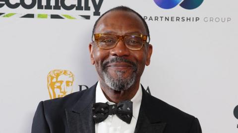 Lenny Henry smiles at the camera as he attends an awards ceremony. He is wearing brown glasses as well as a black suit, bow tie and white shirt.