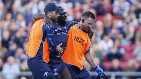 Gabriel Ibitoye (centre) has his arms around the shoulders of two medical staff (left and right) as he is helped walk off the pitch
