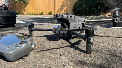 A grey and black drone is seen from the side, while resting on tarmac in a carpark. It has several wings at the end of its legs and has a camera on its main body. Next to the drone is a controller, with a digital screen, several buttons and a joystick. 