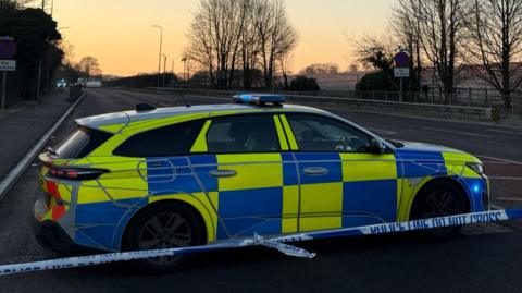 A police car is parked on the A5 on the other side of police tape. The sun is rising.