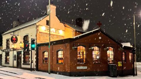 The front of a pub in the snow. Part of the building is white brick and has a large "joules" sign on the side of it. An extension of the building is brown brick with arched windows. Snow is falling from the sky and there is snow on the pavement and nearby road.