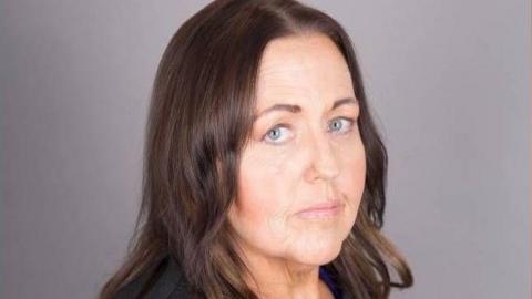 Joanne Monk is pictured standing to the side with her head facing the camera. She has shoulder length brown hair.