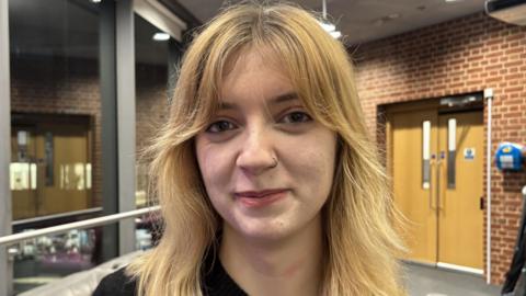 Grace Murray, standing in a room, looking straight at the camera, with long blonde hair and a nose ring on her left side. She is smiling. The building has a door to the right and red bricks. She is wearing a black top. 
