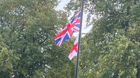 A lamppost with a Union flag and England flag attached, and trees in the background.