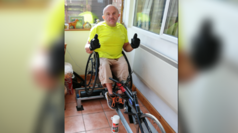 Les Hampton in his sports wheelchair in his conservatory at home, wearing a bright yellow t shirt and sand colour three quarter length cargo trousers. He has trainers on  and black gloves, with both hands in a thumbs up sign to the camera. 