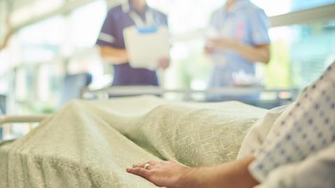 Man with a wedding band on his left hand wearing a hospital gown and lying in a hospital bed. Two nurses can be seen by the bed with a clipboard. 