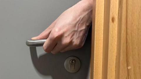 A hand reaching through a partially opened grey door and holding the metal handle. 