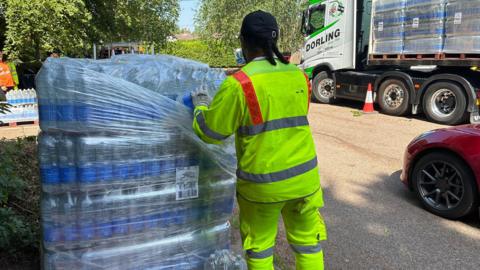 A man in a yellow hi-vis jacket and trousers taking slabs of water out of its shrink wrapping. There is a red car and lorry near him.