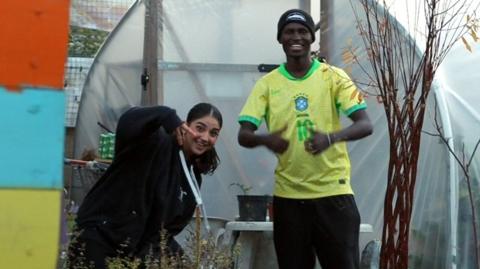 A young woman and a young man smile and make poses in a garden, standing in front of a greenhouse