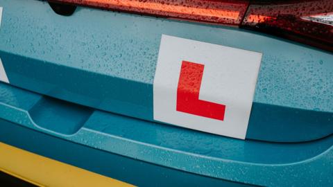 The rear of a teal coloured car showing a learner driver plate attached to the boot.