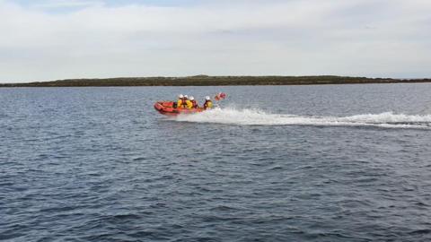 A RNLI lifeboat in the water. Land can be seen behind the boat and white water surrounding the orange boat. Four people are in the boat with yellow jackets, orange life vests and white helmets.