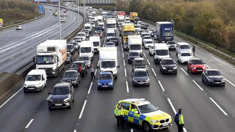 Six lanes of traffic on motorway have all been stopped with a police car parked in the centre. The cars are facing the camera and the image has been taken from  above the M25.